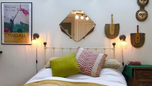 Ein Bett mit weißem Metallgestell, einem grün-rot gestreiften Kissen und einer beigen Decke. An der Wand hängt ein Poster, und über dem Bett befindet sich ein Spiegel.