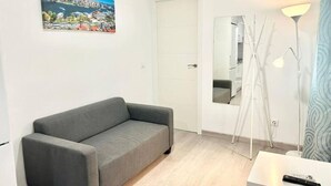 A modern living room with a grey sofa, a painting of a cityscape, a white door, a mirror, a white table with a remote control, and a floor lamp. - Metro Avenida de América 09 (1-bedroom apartment) (Madrid)