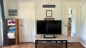 Un salon avec un téléviseur posé sur un meuble, une porte avec une pancarte et une étagère où sont pliées des serviettes.