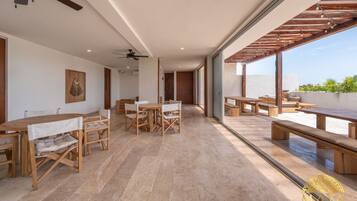 A spacious indoor area with wooden tables and chairs, a ceiling fan, and large windows offering a view of the outdoors.