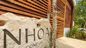 A stone sign with "NHOA CONDOS" written on it, placed in front of a wooden building with slatted roof design. - NHOA 303C Lux 1BR Condo Pool View in Aldea Zama (Tulum)