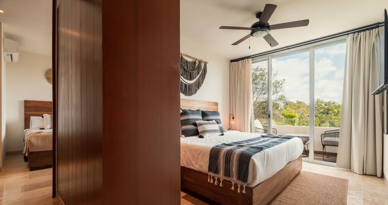 Une chambre dotée d’un grand lit, d’un ventilateur de plafond et offrant une vue sur l’extérieur grâce à de grandes fenêtres.