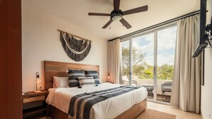 A bedroom with a large bed, a ceiling fan, a nightstand, a wall-mounted lamp, and a view of the outdoors through a sliding glass door.