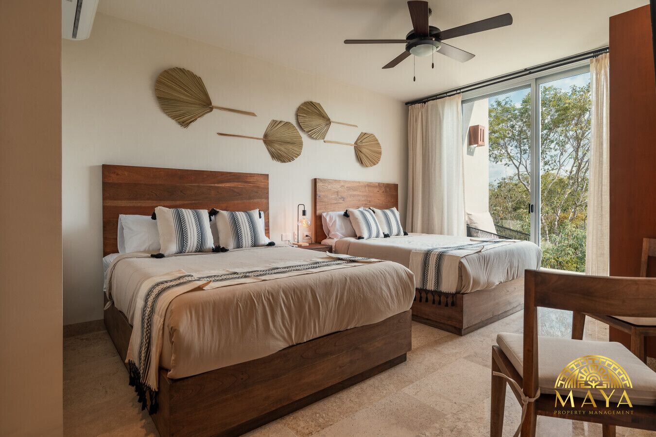A hotel room with two beds, a ceiling fan, and a large window with a view of trees.