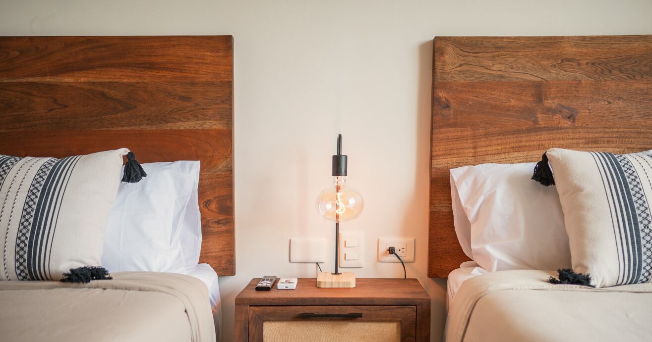 A hotel room with two beds, a nightstand with a lamp, and a small table.