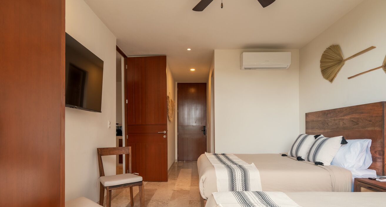 A hotel room with two beds, a TV mounted on the wall, a wooden chair, and a door leading to a hallway.
