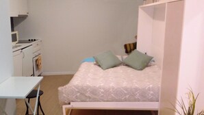 A small, neatly organized bedroom with a single bed, two pillows, a white desk, and a small kitchenette.