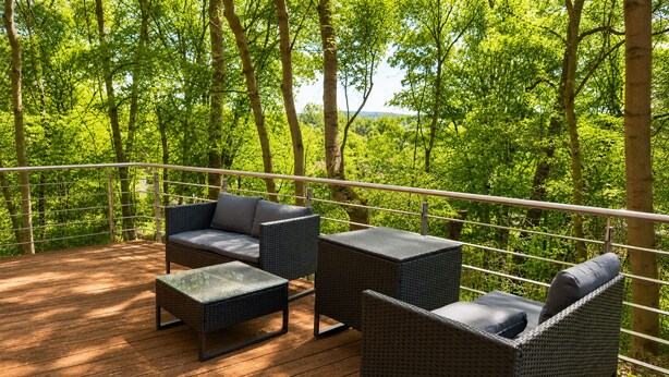 En udendørs terrasse med fletningsmøbler og et glasbord, omgivet af frodig vegetation.