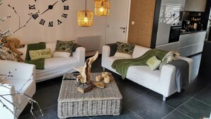 A modern living room with two white sofas, a large wall clock, and a kitchen area in the background. - Cottage in Houyet Near Lesse River Trails (Houyet)