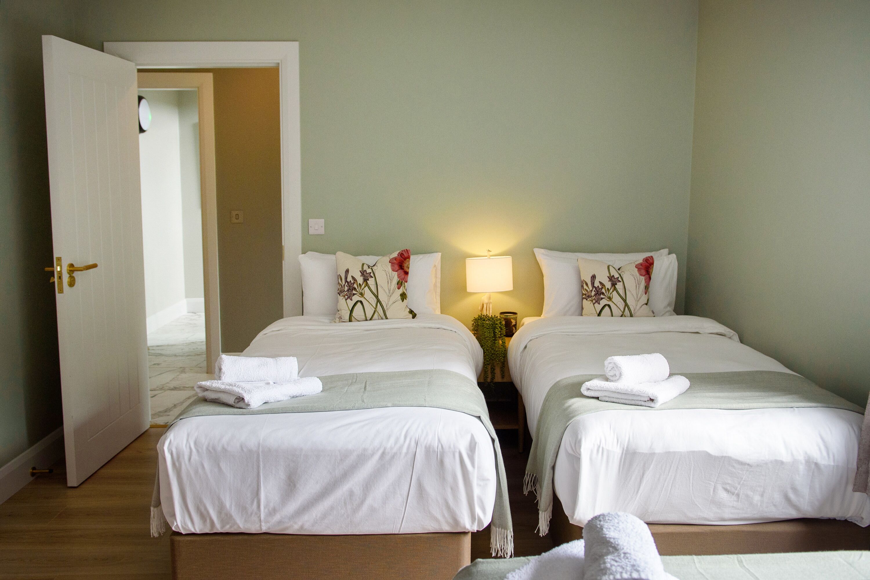 Une chambre d’hôtel avec deux lits, une porte, une table de chevet avec une lampe et une petite table avec une plante.