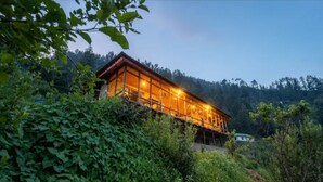A wooden cabin with a glass facade, surrounded by dense greenery and hills in the background.