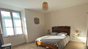 Un dormitorio con piso de madera, una cama con cubrecama gris, una mesita de noche de madera con una lámpara, una silla y una ventana con persianas.