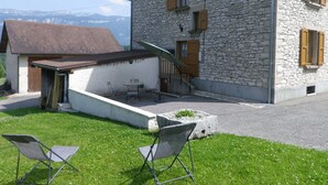 Un edificio de piedra con techo inclinado, una estructura pequeña adosada con puerta de madera y un área ajardinada con dos sillas.