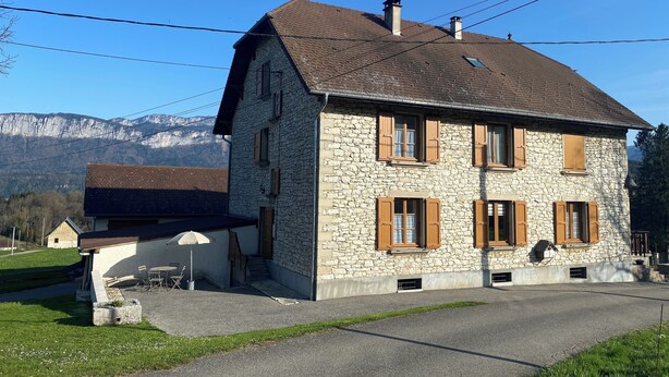 Una casa de piedra con techo inclinado, contraventanas de madera y un pequeño espacio al aire libre con mesa y sillas.