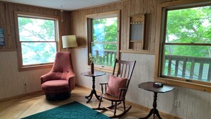 A room with wooden walls, a rocking chair, a patterned armchair, a small table, and a green area rug. - Charming Vintage Lakefront Cottage on Seneca Lake (Penn Yan)
