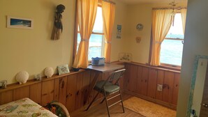 A room with wooden walls, a desk with a chair, a bed with a floral bedspread, and a view of the sea through windows with yellow curtains. - Charming Vintage Lakefront Cottage on Seneca Lake (Penn Yan)