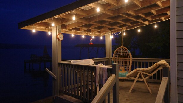 A wooden deck with a hanging chair and a swing, string lights, and a clock. - Charming Vintage Lakefront Cottage on Seneca Lake (Penn Yan)