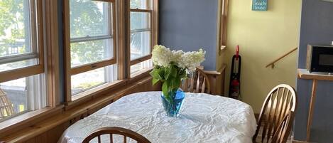 Un comedor con una mesa redonda cubierta con mantel blanco, rodeada de sillas de madera. Un jarrón con flores blancas está sobre la mesa.