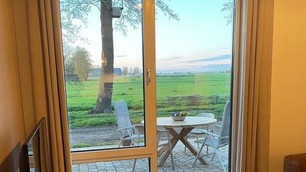 A room with a large sliding glass door leading to an outdoor patio with a table and chairs, a view of a green field, and a tree. - Apartment in Workum Near Ijsselmeer Beach (Workum)
