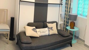 A modern living room with a grey sofa, two decorative pillows, a small round table with a lamp, and a window with curtains.