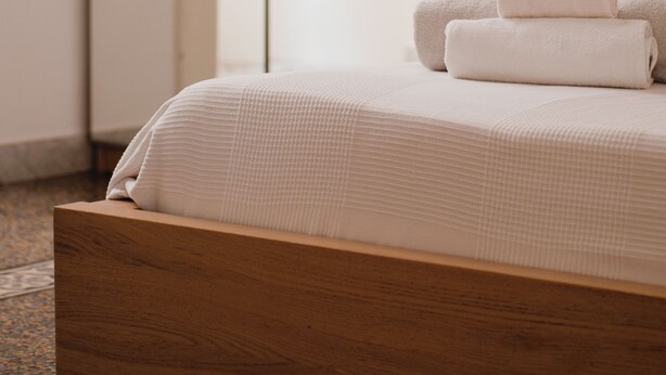 Una cama bien hecha con cabecera de madera, un ventanal con vistas a plantas en macetas y una puerta corredera de vidrio.