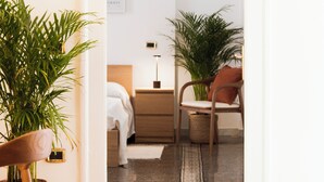 Habitación de hotel con cama, mesita de noche, lámpara, sillón y plantas en macetas.