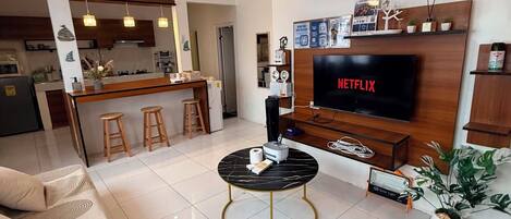 A modern living room with a flat-screen TV, a dining area with bar stools, and a kitchen in the background.