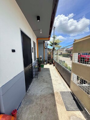 A narrow outdoor corridor with a concrete floor, a metal gate, and a small garden area with potted plants.