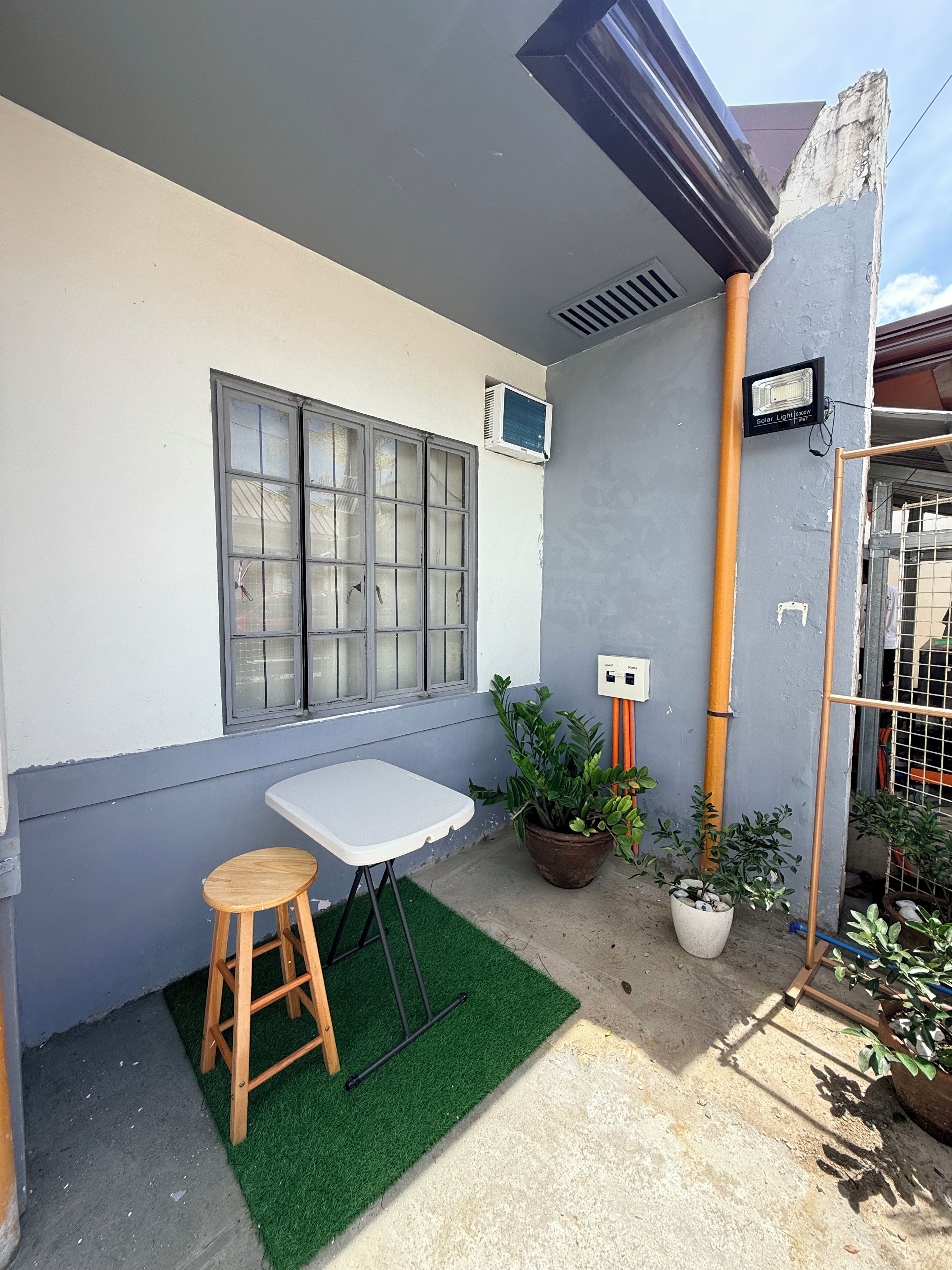 Un pequeño patio al aire libre con una mesa, un taburete, plantas en macetas y una ventana con unidad de aire acondicionado.