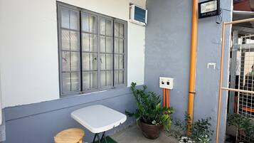 Un pequeño patio al aire libre con una mesa, un taburete, plantas en macetas y una ventana con unidad de aire acondicionado.