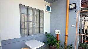 Un petit espace extérieur aménagé avec une table et un tabouret, entouré de plantes en pot et d’un mur gris.
