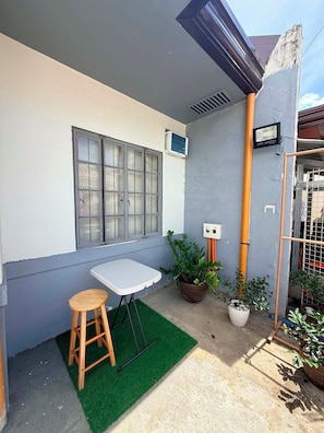 A small outdoor seating area with a table and stool, surrounded by potted plants and a grey wall.