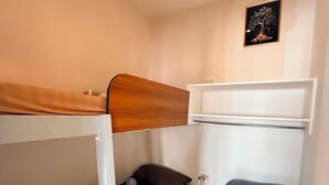 Une petite chambre avec un lit superposé, une petite table de chevet avec une lampe et une étagère fixée au mur.