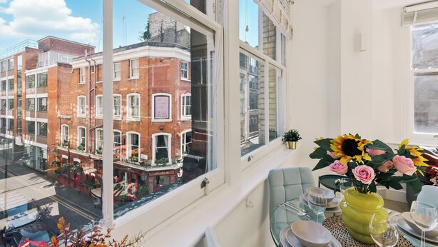 Una habitación con un ventanal grande que da a la calle, una mesa de comedor con sillas y un jarrón con flores.