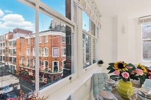 A room with a large window overlooking a street, a dining table with chairs, and a vase with flowers.