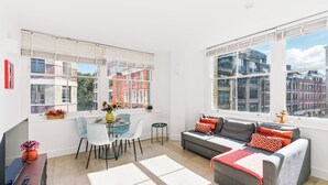 Una sala de estar moderna con un sofá gris, sillas blancas y azules, una mesa de comedor y ventanas amplias con vistas a la ciudad.