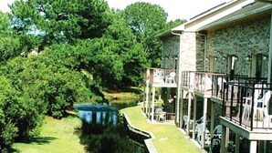 A riverside property with a stone building, a covered patio with outdoor seating, and lush greenery.