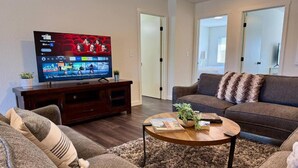 A living room with a flat-screen TV, a dark wood entertainment unit, a round coffee table with books and a plant, and a grey sofa with a textured throw. - Homestead Village - Two Bedroom Condo Top Floor (Sandpoint)