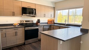 A modern kitchen with wooden cabinets, stainless steel appliances, and a large window providing a view of trees. - Homestead Village - Two Bedroom Condo Top Floor (Sandpoint)