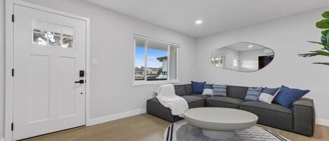 A modern living room with a grey sectional sofa, a round coffee table, a large mirror, and a view of the outside through a window.
