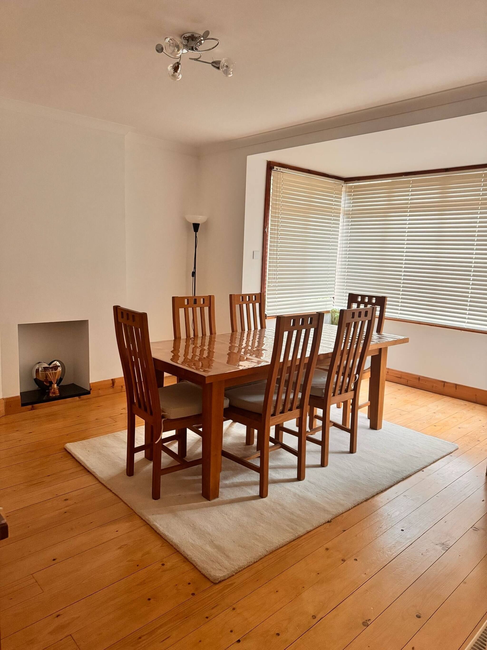 A dining area with a wooden table and chairs, a chandelier, a floor lamp, and a large window with blinds.