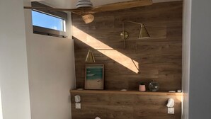 A bedroom with a wooden headboard, a bed with white bedding, a ceiling fan, a small window, and wall-mounted lights.