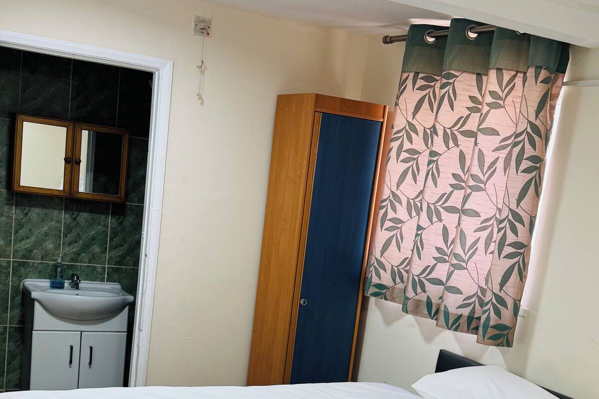 Ein ordentlich gemachtes Bett mit weißer Bettwäsche, eine Holztür, ein Badezimmer mit Waschbecken und Spiegel sowie ein Fenster mit einem vorblauen Vorhang.