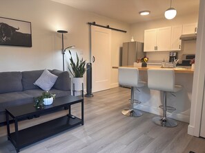 Un salon moderne avec un canapé, une table basse et une plante. À côté se trouve une cuisine avec des meubles blancs, un tabouret de bar et une machine à café.