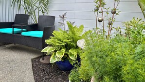 Un'area patio con un divano in rattan nero e cuscini color turchese, una pianta in vaso grande e altra vegetazione.