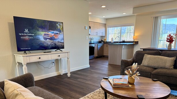 A modern living room with a flat-screen TV, a sofa, a coffee table, and a kitchen in the background. - Homestead Village - Two Bedroom Condo Top Floor (Sandpoint)