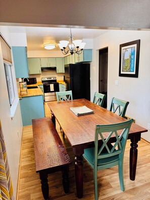 Een eetgedeelte met een houten tafel en stoelen, een bank, een kroonluchter, en een keuken op de achtergrond.