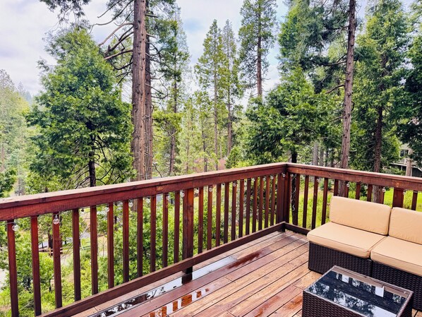 A wooden deck with a sofa and a glass-top table, surrounded by tall trees.