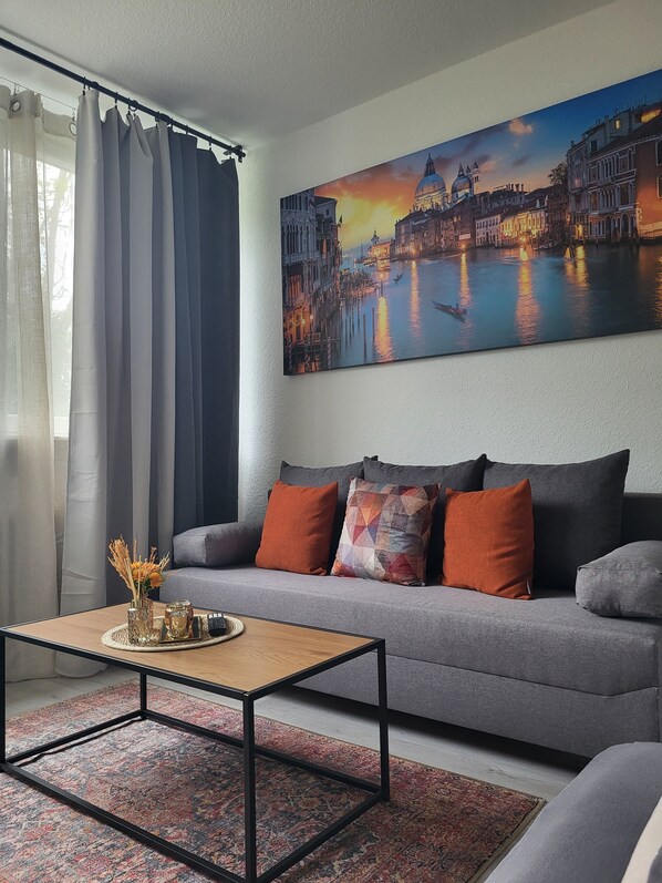 A living room with a grey sofa, a coffee table, and a large painting of a cityscape. - Charming & cozy apartment in Wuppertal incl. tea & coffee (Wuppertal)