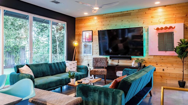 A modern living room with a large TV, green sofas, a wooden wall, and a ceiling fan. - Modern ATX Mansion Walk to 6th Street (Austin)